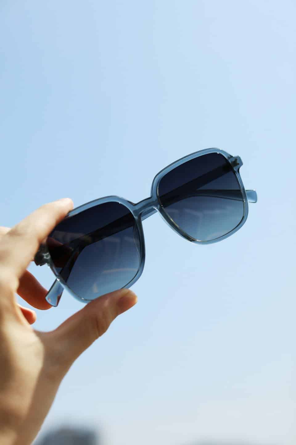 A hand holds blue-tinted square sunglasses against a clear sky.