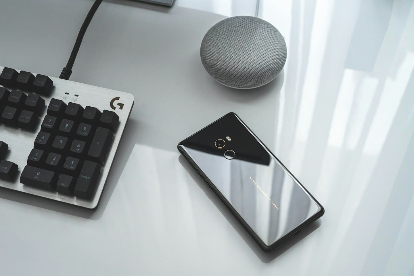 Smartphone, keyboard, and smart speaker on a white desk.